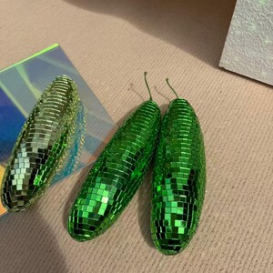 May include: Three green disco ball-style decorative objects. Two are elongated, with a stem-like detail at the top. The third is a similar shape, but smaller. All are covered in small, reflective squares, creating a shimmering effect.