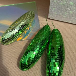 May include: Three decorative disco ball cucumbers. Two are bright green, and one is a lighter green. The cucumbers are covered in small, reflective squares, creating a shimmering effect. The cucumbers have small green stems.