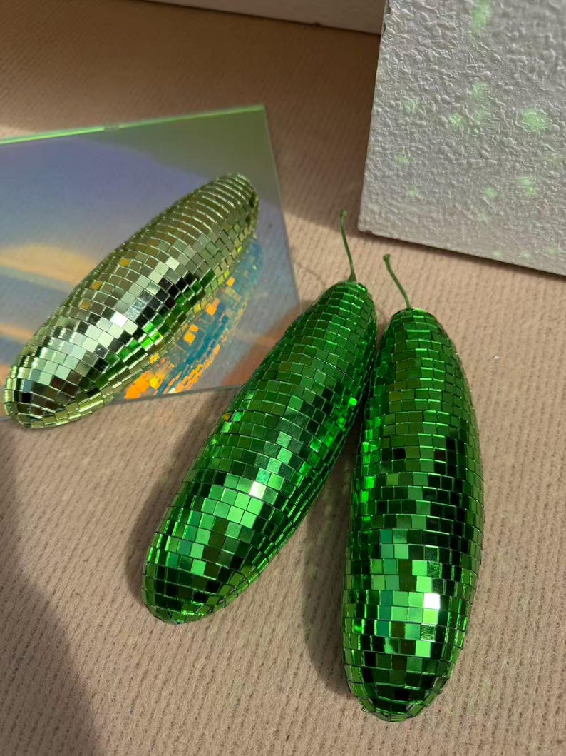 May include: Three decorative disco ball cucumbers. Two are bright green, and one is a lighter green. The cucumbers are covered in small, reflective squares, creating a shimmering effect. The cucumbers have small green stems.