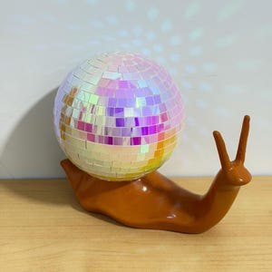 Puede incluir: Una caprichosa figura de caracol de cerámica con una concha de bola de discoteca. El caracol es de color marrón cálido, y la concha está cubierta de azulejos iridiscentes y reflectantes. La figura está sobre una superficie de madera.
