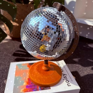 May include: A mirrored disco ball globe, mounted on a brown stand and an orange base. The globe sits on a book with the words "noisy crowd" on the cover. The globe is a fun decorative item.