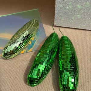 May include: Three green, pear-shaped ornaments with a disco ball effect. One lies on a reflective surface, two stand upright. Each is covered in small, square, mirrored tiles. The background is a neutral beige colour.