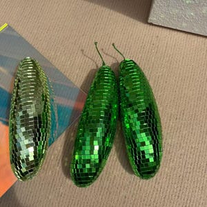 May include: Three green disco ball-style ornaments shaped like pickles. Each pickle is covered in small, reflective squares. Two pickles have green stems. The ornaments are on a neutral-coloured surface.