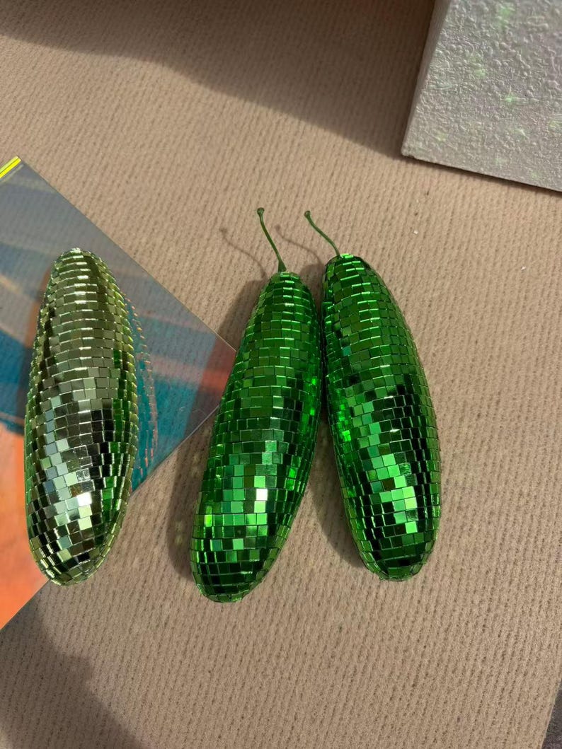 May include: Three green disco ball-style ornaments shaped like pickles. Each pickle is covered in small, reflective squares. Two pickles have green stems. The ornaments are on a neutral-coloured surface.