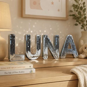 Op de afbeelding: Vier vrijstaande letters die het woord LUNA vormen, elk bedekt met kleine, reflecterende zilveren spiegeltegels. De letters hebben een blokstijl lettertype en een driedimensionale vorm, wat een mozaïekeffect creëert dat licht in de omgeving weerkaatst.