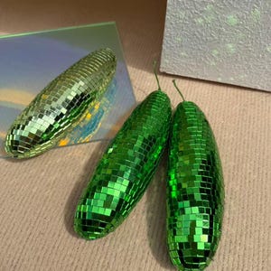 May include: Three decorative disco ball-style objects. Two are bright green, elongated, and have small square mirrored tiles. The third is a similar shape, but with a gold-green hue. The objects are arranged on a neutral surface.