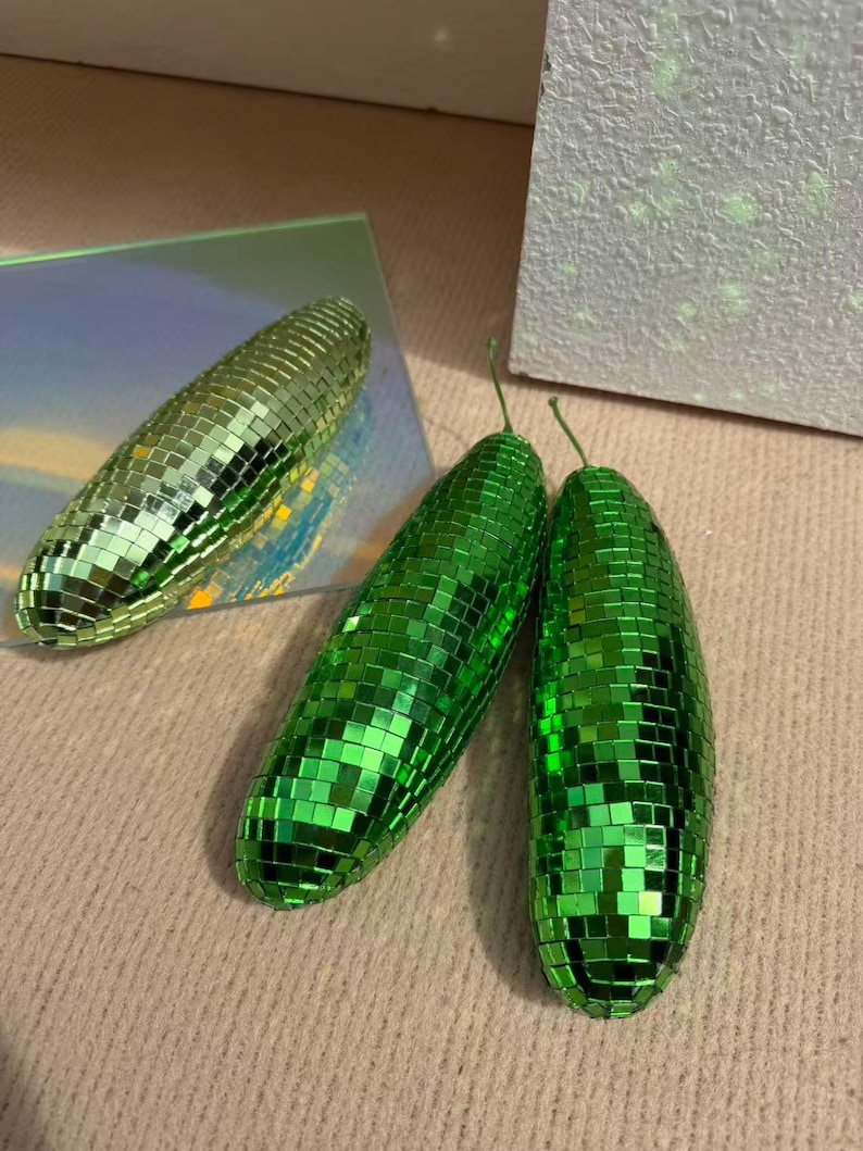 May include: Three decorative disco ball-style objects. Two are bright green, elongated, and have small square mirrored tiles. The third is a similar shape, but with a gold-green hue. The objects are arranged on a neutral surface.