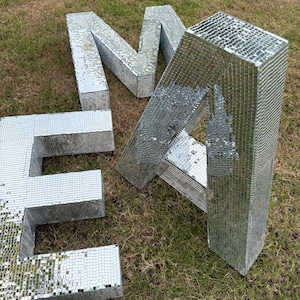 Puede incluir: Tres letras tridimensionales grandes que forman la palabra EMA, cada una cubierta con pequeñas piezas de espejo plateadas y reflectantes. Las letras son independientes y tienen una fuente de estilo bloque, creando un efecto de mosaico.
