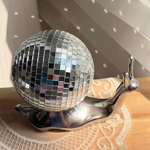 Puede incluir: Una bola de discoteca plateada se asienta sobre una figura decorativa de caracol. La bola está cubierta de pequeños cuadrados reflectantes. La figura del caracol es de metal plateado y descansa sobre un tapete crema con dibujos.