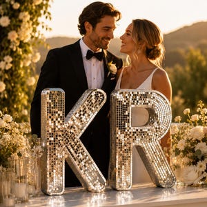 May include: Two large, three-dimensional letters K and R covered in small, reflective silver mirror tiles. The letters are positioned on a white surface, creating a sparkling, grid-like surface texture that reflects light from the surrounding environment.