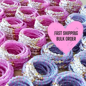 May include: An assortment of beaded bracelets in shades of pink, white, and blue. The bracelets feature white letter beads and gold accents. The image includes a pink heart with the text "FAST SHIPPING BULK ORDER".
