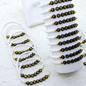 May include: White beaded bracelets with black and gold letter beads spelling "FREEDOM". The bracelets are displayed on a white surface and a white cylindrical object. Gold beads separate the letter beads.