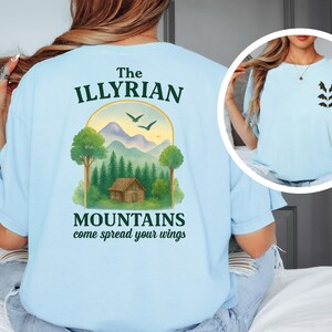 May include: Light blue t-shirt with a graphic on the back featuring a mountain scene with a cabin, trees, and birds, and the text "The Illyrian Mountains come spread your wings." A smaller image on the front shows bat silhouettes.