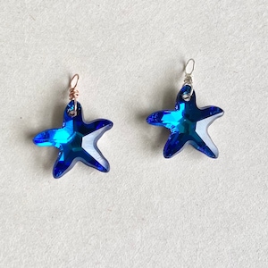 May include: Two blue crystal starfish pendants. One has a copper-colored wire loop, and the other has a silver-colored wire loop. The starfish have a faceted design that reflects light, creating a sparkling effect. The pendants are likely for jewelry making.