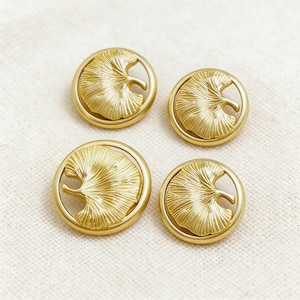 May include: Four gold-colored buttons with a ginkgo leaf design. Each button features a detailed leaf within a circular frame. The buttons are arranged on a light-colored fabric, showcasing their metallic sheen and intricate design.