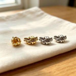 May include: Four small metal stud earrings arranged in a row. The first is a gold-toned flower-shaped stud, followed by three knot-shaped studs, with one gold-toned knot and two silver-toned knots, all featuring a polished, reflective metallic finish.