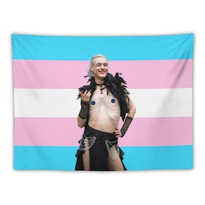 May include: A tapestry displaying the transgender pride flag in blue, pink, and white. In front of the flag, a person is wearing a black feather boa, a black choker, and a black skirt with chain details. The person is also wearing blue nipple pasties.