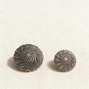 Puede incluir: Dos botones de metal plateado con un diseño en espiral. El botón más grande tiene un diámetro de aproximadamente 3,2 cm, mientras que el más pequeño mide alrededor de 1,9 cm. Ambos botones tienen un borde texturizado y una hendidura central circular.