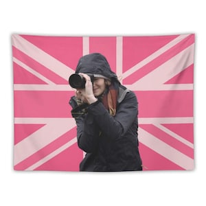 May include: A pink and white Union Jack flag tapestry with a person holding a camera. The person is wearing a black jacket and a hood. The flag is a vibrant pink with white stripes.