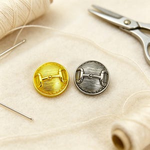May include: Two round buttons, one gold-toned and one silver-toned, each featuring a textured surface and a central horizontal metal bar detail. The buttons are positioned near sewing supplies including a spool of thread and metal scissors.