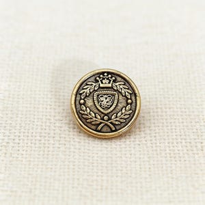 Peut inclure: Un bouton rond de couleur dorée avec un motif de blason détaillé. Le blason comprend une couronne, un écusson avec des lettres ornées et est entouré de feuilles de laurier. Le bouton est posé sur un fond texturé blanc cassé.