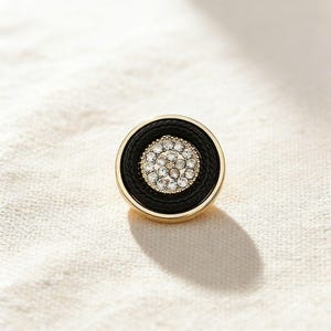 Op de afbeelding: Een ronde knoop met een zwart midden, een goudkleurige rand en een ring van heldere strass-steentjes. De knoop heeft een getextureerde zwarte rand en een goudkleurige buitenrand. De knoop is geplaatst tegen een witte achtergrond.