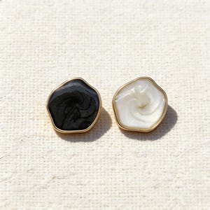 Puede incluir: Un par de pendientes de botón con formas irregulares y orgánicas. Un pendiente es negro con un patrón arremolinado, el otro es blanco con un diseño similar. Ambos están engastados en marcos de metal color oro, creando un aspecto contrastante y elegante.