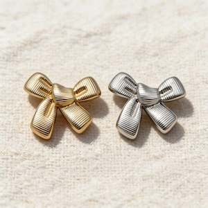 May include: Two bow-shaped accessories. One is gold-colored, the other is silver-colored. Both have a ribbed texture and are set against a neutral, textured background. These could be used as jewelry or decorative accents.