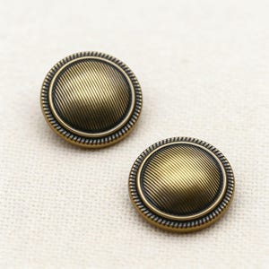 Peut inclure: Deux boutons de couleur laiton antique avec un motif strié. Chaque bouton a une forme circulaire avec un centre texturé et un bord extérieur décoratif. Les boutons sont posés sur un fond neutre.