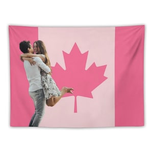 Op de afbeelding: Een roze wandtapijt met het Canadese vlagontwerp, inclusief een groot esdoornblad. Een stel omhelst elkaar voor de vlag. Het rechthoekige wandtapijt is gemaakt van stof.