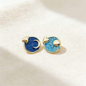 May include: A pair of celestial stud earrings. One earring features a dark blue crescent moon design, the other a light blue sun with golden accents. Both earrings have a gold-coloured border and post.