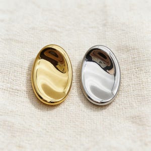 May include: Two oval-shaped, polished metal objects. One is gold-toned, the other is silver-toned. Both have a reflective surface, and are set against a neutral, textured background.