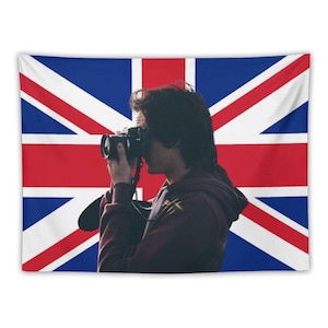 Puede incluir: Un tapiz con la bandera del Reino Unido, la Union Jack. Una persona sostiene una cámara frente a la bandera. La bandera tiene un fondo azul con rayas rojas y blancas.