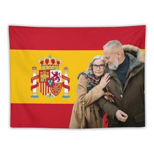 Könnte beinhalten: Ein Wandteppich mit der spanischen Flagge und einem sich umarmenden Paar davor. Die Flagge ist rot und gelb, mit dem spanischen Wappen. Das Paar trägt Mäntel und Schals.
