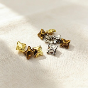 Op de afbeelding: Een verzameling stervormige metalen studs in goud-, zilver- en bronskleuren. De studs zijn verspreid op een witte, getextureerde ondergrond, met zachte belichting die highlights en schaduwen creëert. De studs hebben een vierpuntig sterontwerp.