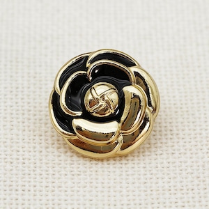 May include: A round button shaped like a camellia flower, featuring a polished gold-tone metal frame with smooth, glossy black enamel inlay filling the petals and a small, textured gold-tone circular center piece with a cross-hatch design.
