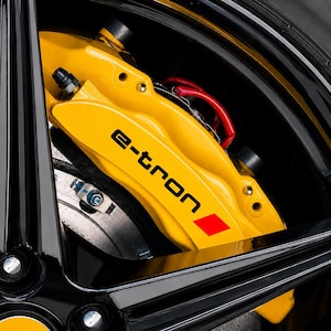 Peut inclure: Un étrier de frein automobile jaune vif portant l'inscription noire e-tron et un petit logo carré rouge. L'étrier est monté derrière une jante noire brillante, avec un disque de frein métallique et une durite de frein rouge en arrière-plan.