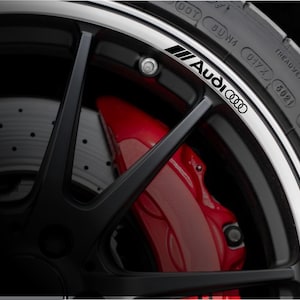 May include: Close-up of a black car wheel with a silver rim. The wheel features a red brake caliper and the Audi logo in black. The tire has white lettering with the brand and size.