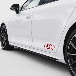 Può includere: Un'auto bianca con un adesivo rosso del logo a quattro anelli Audi sul pannello laterale inferiore. Il veicolo presenta cerchi in lega scuri a razze multiple, un alloggiamento dello specchietto laterale nero e un design della carrozzeria elegante.