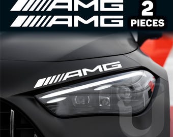 May include: White AMG car decals. The image shows two sets of AMG decals, with the text "2 PIECES" in a white box. One set is placed on the hood of a black car, near the headlight.