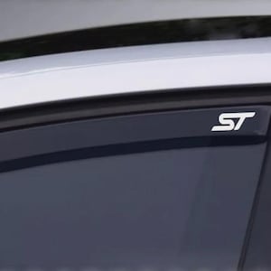 Op de afbeelding: Een close-up van een zijruit van een auto met een wit "ST"-embleem. De ruit is getint en het dak van de auto is zichtbaar op de achtergrond. De auto lijkt zilverkleurig of licht van kleur.