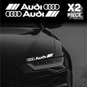 May include: White Audi car decal set. The image shows a black car with white decals. The decals include the Audi logo and the word "Audi" in a stylized font, along with the text "X2 PIECE".