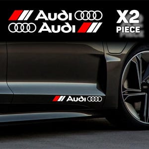 以下が含まれることがあります： 白と赤のアウディ車のデカール。デカールにはアウディのロゴと「Audi」の文字が白で、赤いストライプが入っています。画像は、黒い車にデカールが貼られている様子で、「X2 PIECE」の文字があります。