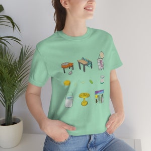 May include: A light green t-shirt with a graphic design of various pottery tools and supplies. The design includes a pottery wheel, a table with a pottery tool, a jar of glaze, a sponge, a pair of scissors, a bottle of water, a small table, and a package of pottery tools.