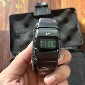 May include: A black digital watch with the Nike logo displayed. The watch face shows the word "TIME" in green. The watch is held in a hand, and is in a black case.
