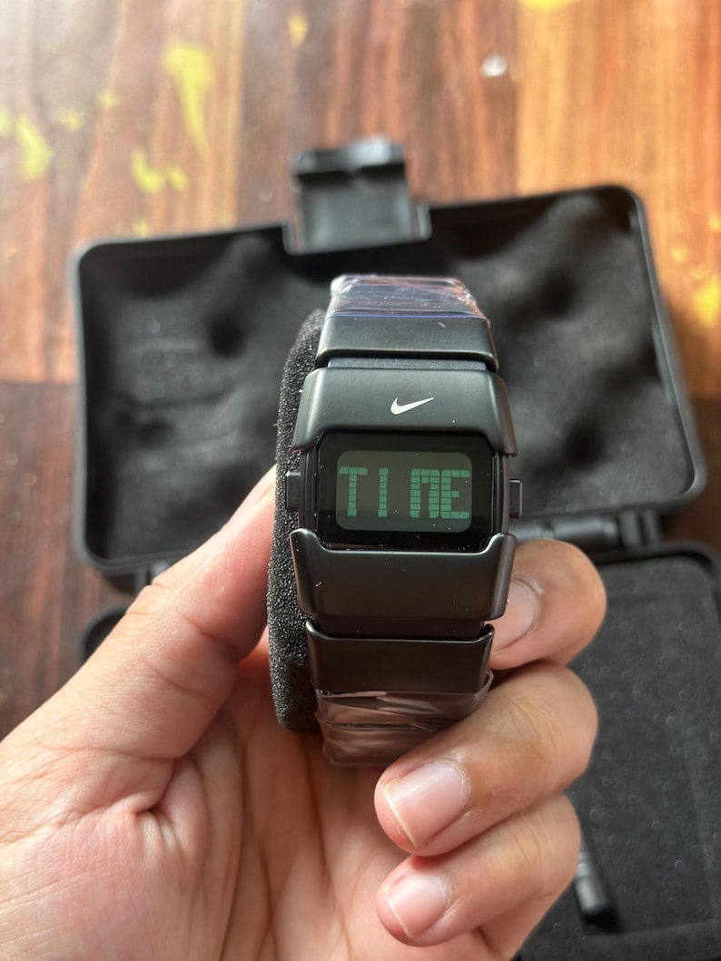May include: A black digital watch with the Nike logo displayed. The watch face shows the word "TIME" in green. The watch is held in a hand, and is in a black case.
