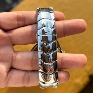 May include: A silver-toned watch band with a unique, textured design. The band features a series of curved, interlocking segments that create a sleek, modern look. The watch band is held in a hand, showcasing its polished, reflective surface.