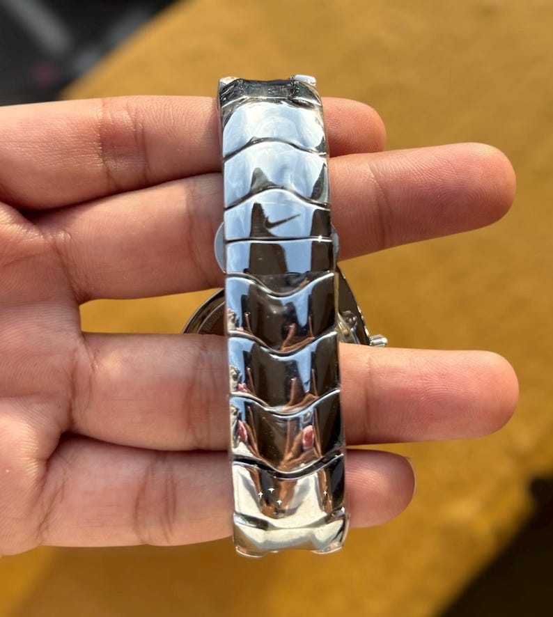 May include: A silver-toned watch band with a unique, textured design. The band features a series of curved, interlocking segments that create a sleek, modern look. The watch band is held in a hand, showcasing its polished, reflective surface.