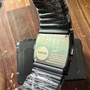 May include: Black digital watch with a rectangular face and a black band. The watch face has text that reads "N20 RESIST 7BAR PERFORMANCE POLYMERS/METALS MINERAL CRYSTAL DESIGNED IN USA". The watch is wrapped in clear plastic.