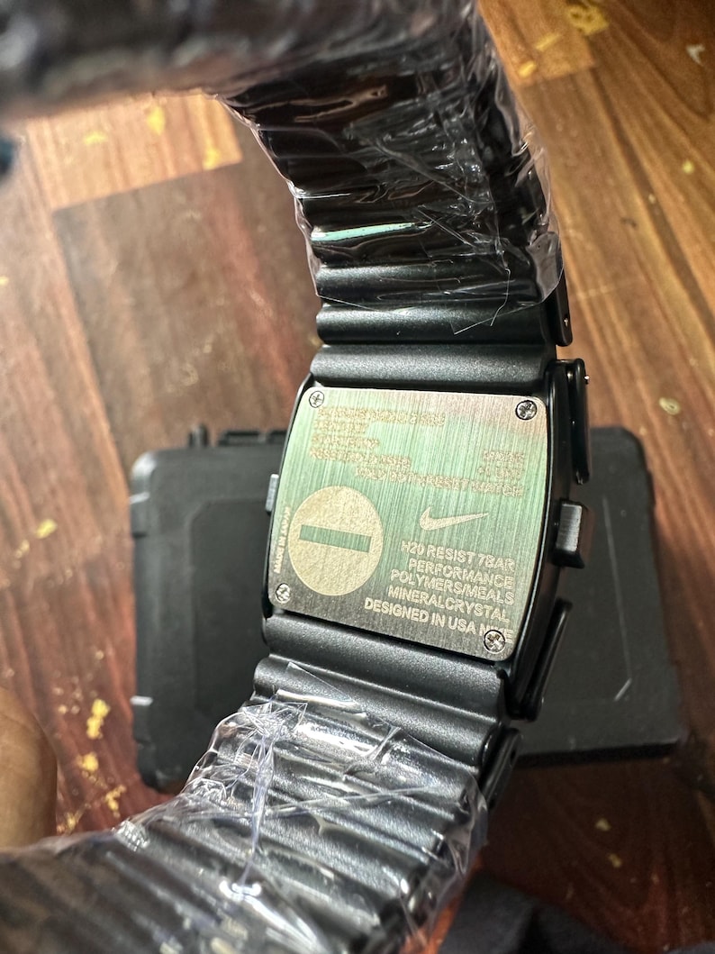 May include: Black digital watch with a rectangular face and a black band. The watch face has text that reads "N20 RESIST 7BAR PERFORMANCE POLYMERS/METALS MINERAL CRYSTAL DESIGNED IN USA". The watch is wrapped in clear plastic.